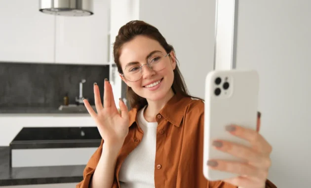 Portrait of stylish beautiful woman at home, video chats on smartphone, connects to online meeting from her living room, lifestyle social media blogger records a video on mobile phone.